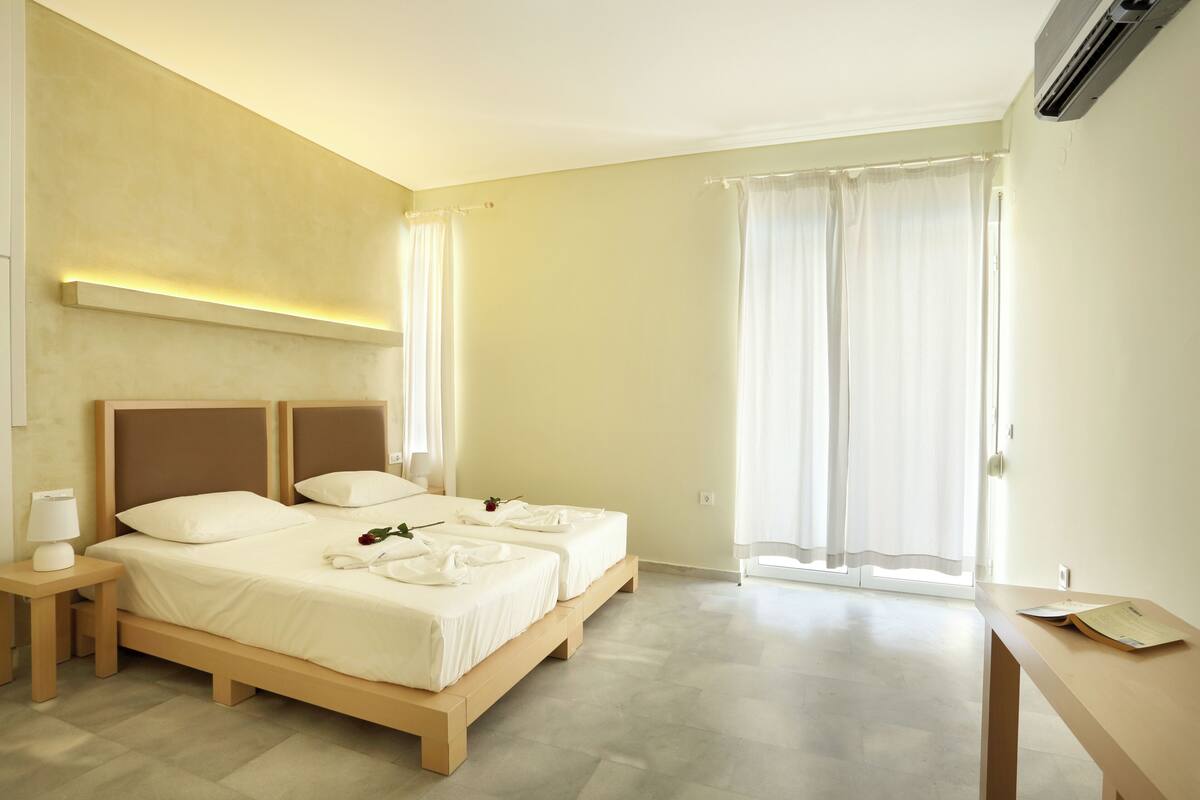 A hotel room with two beds, a bedside table with a lamp, a desk with a chair, and a large window with curtains.