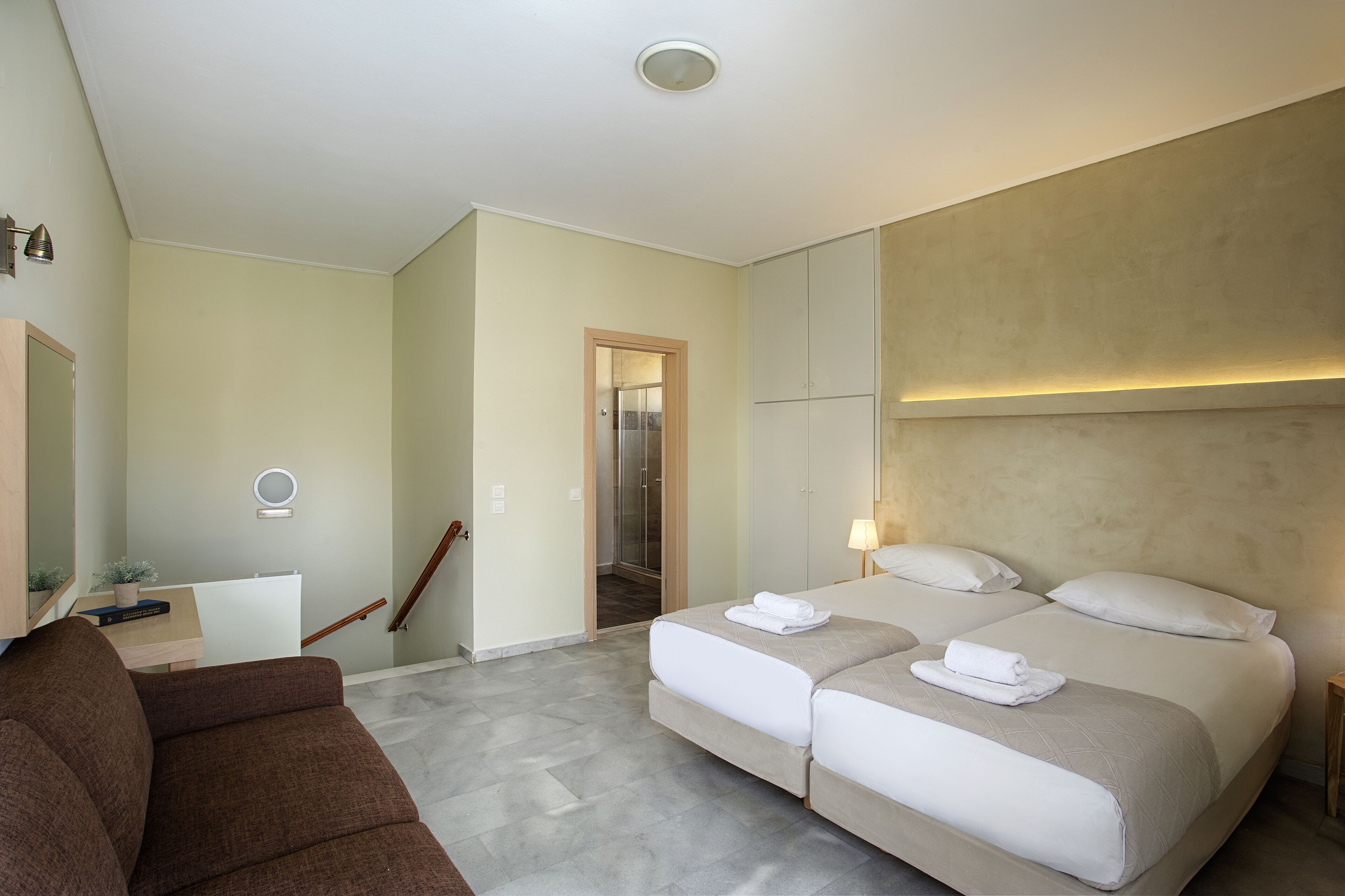 A modern hotel room with a sofa, two beds, a desk, and a shower area.