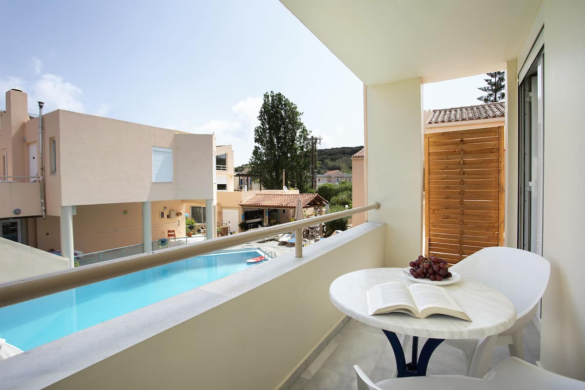 A balcony with a pool, a table with a book and grapes, and a view of buildings and trees.