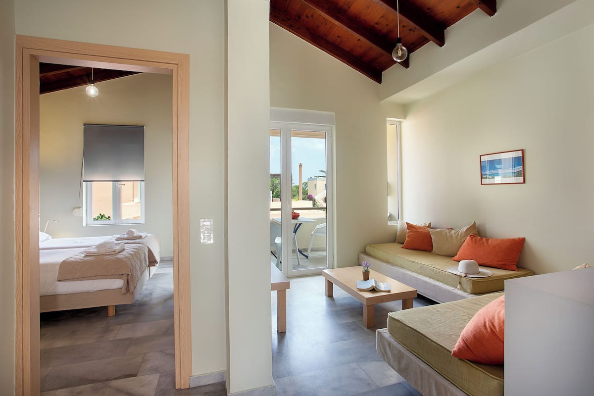 A modern hotel room with a bed, a sofa, a dining table, and a view through a glass door to the outside.