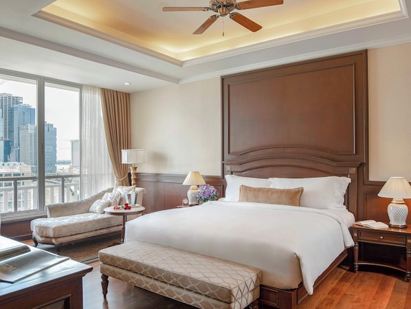 A hotel room with a large bed, a sitting area with a sofa and armchair, a desk, and a view of the city through large windows.