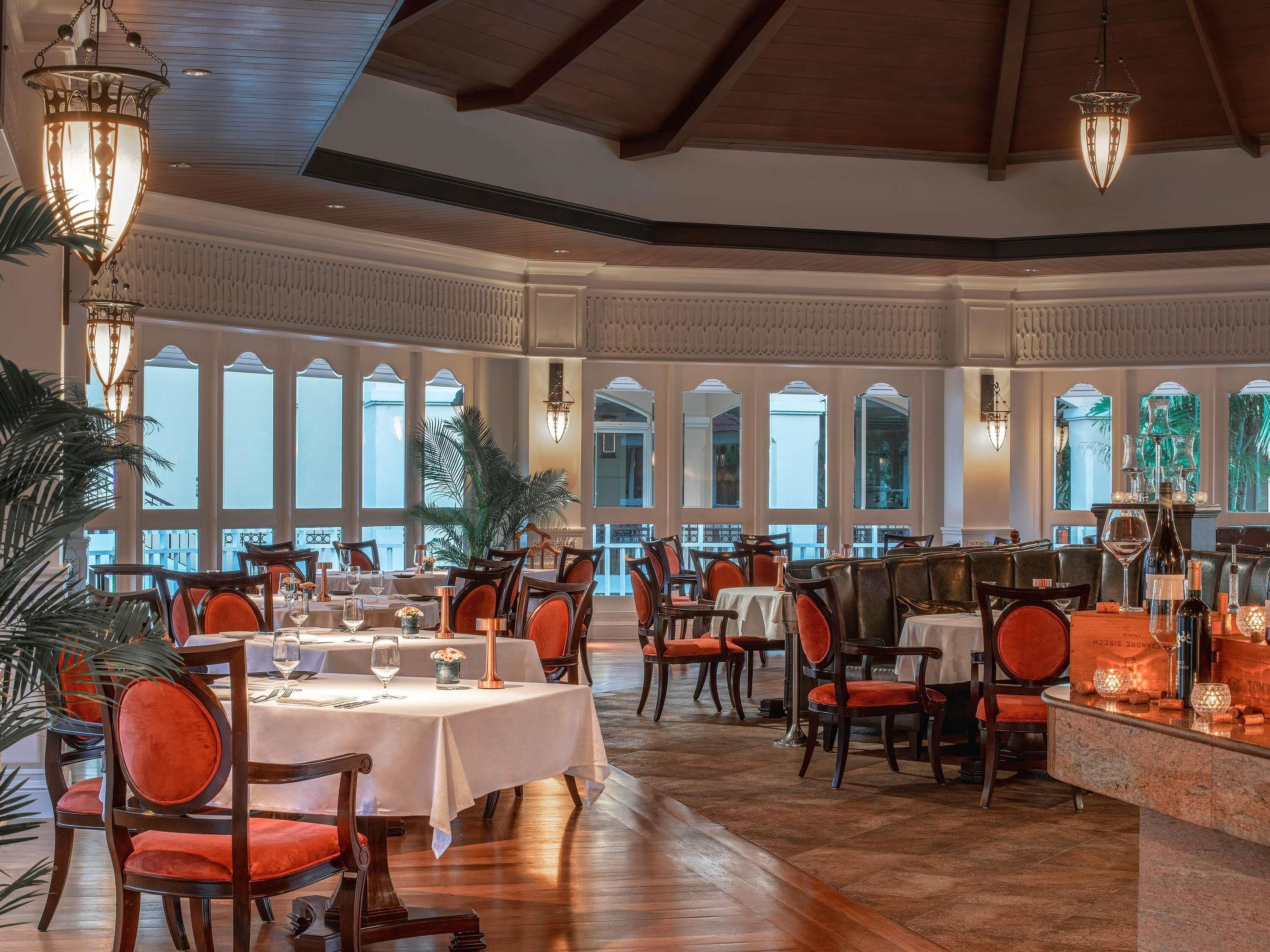 A spacious dining area with multiple tables and chairs, large windows, and decorative lighting fixtures.