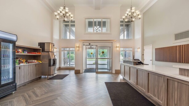 A spacious hotel lobby with a herringbone wood floor, a central glass door entrance, and a reception desk with a wooden backdrop. - Velora Hinton, an Ascend Collection Hotel (Hinton)