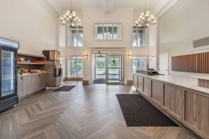 A spacious hotel lobby with a herringbone wood floor, a central glass door entrance, and a reception desk with a wooden backdrop.