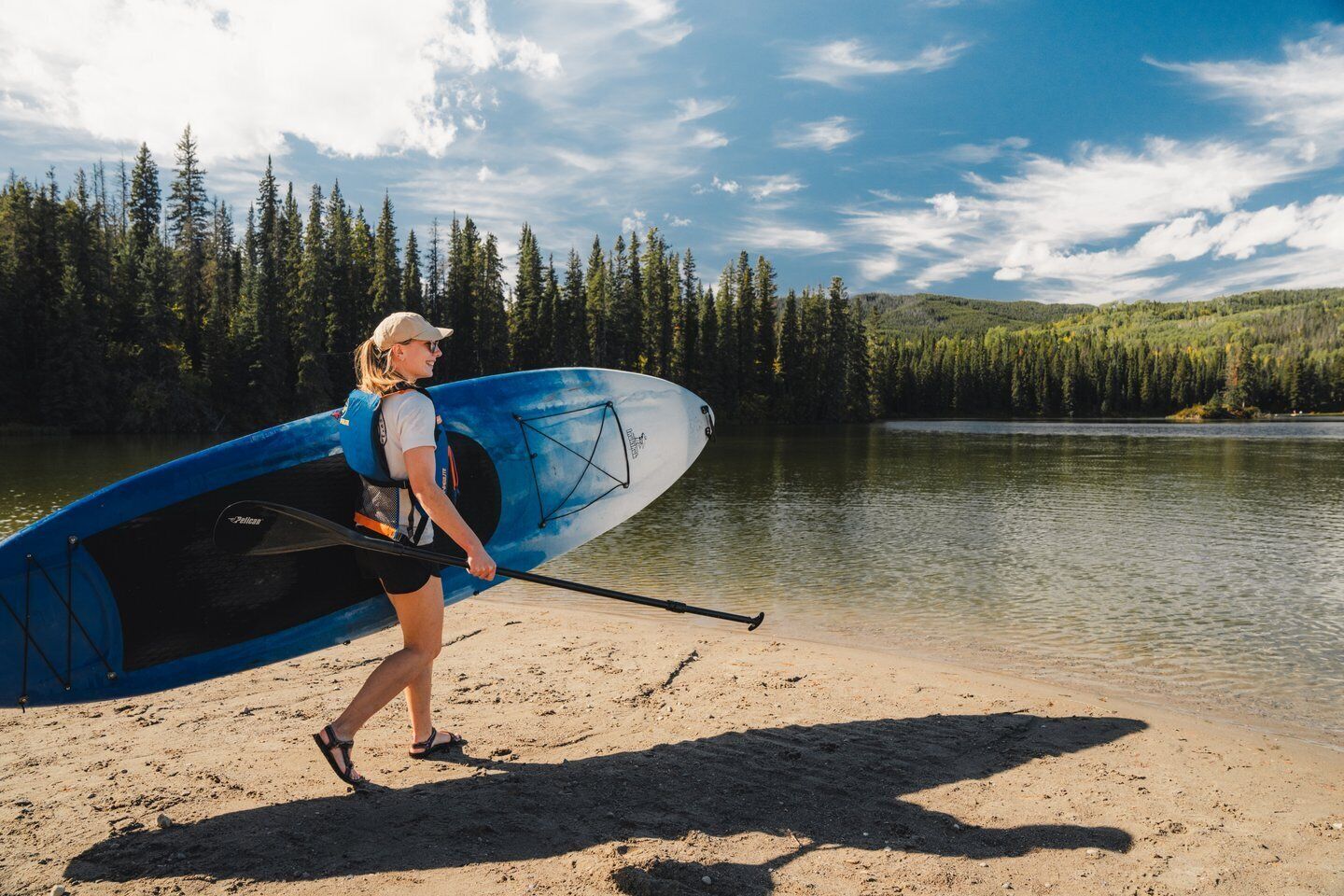 Eine Person trägt ein Stand-Up-Paddleboard an einem sandigen Strand, im Hintergrund ein von Bäumen gesäumter See.