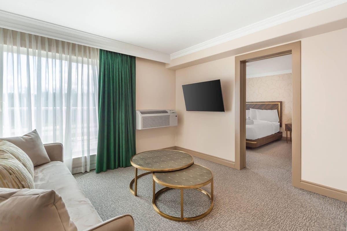 A hotel room with a sofa, two round tables, a flat-screen TV, an air conditioning unit, and a bed with a headboard.