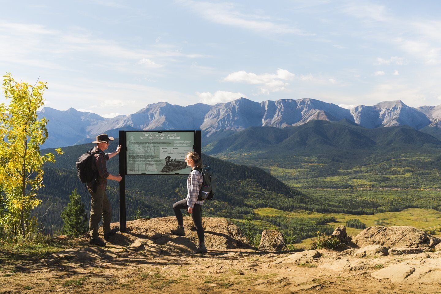A mountain view with a signboard and two people standing beside it.