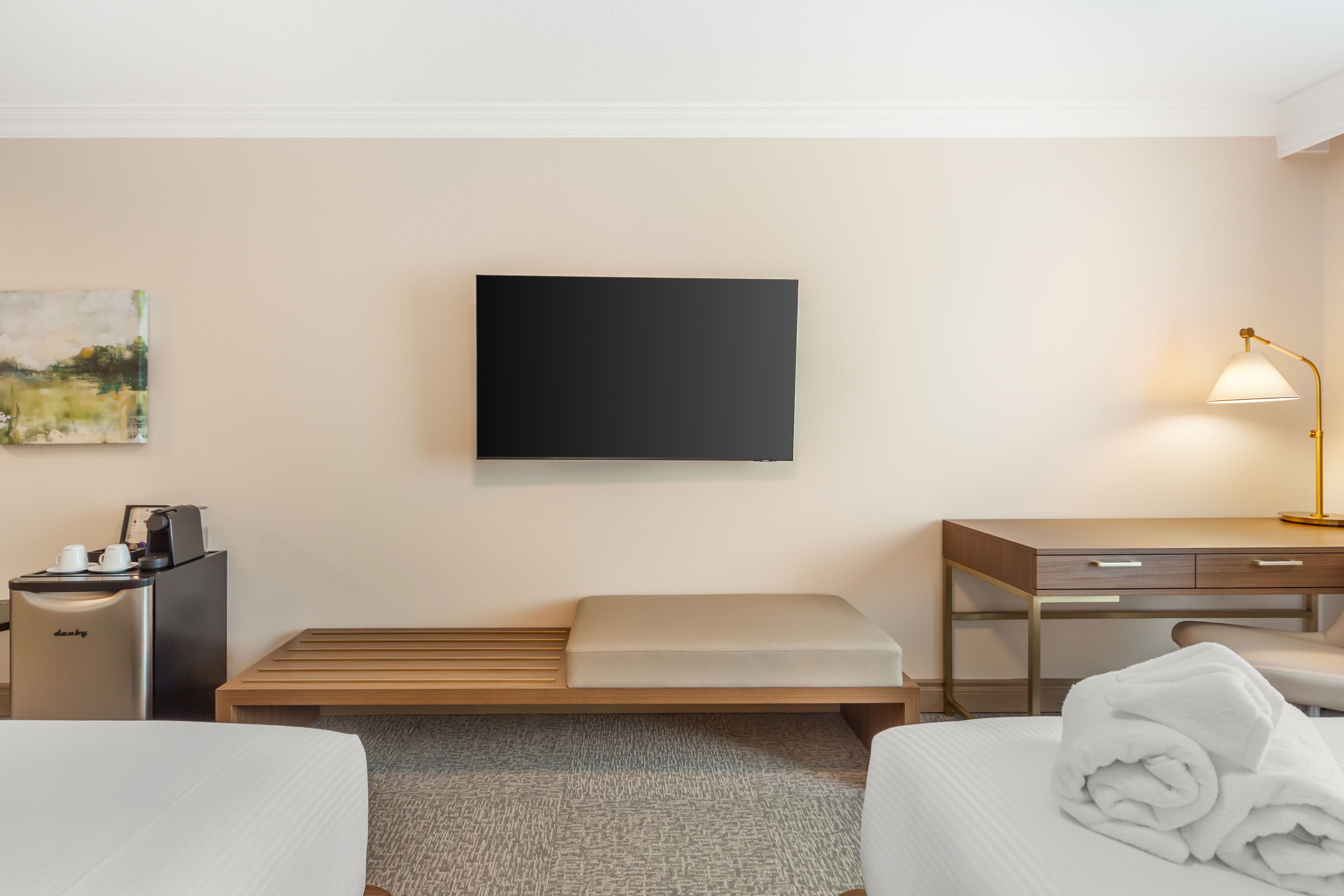 A hotel room with a bed, a desk, a chair, a lamp, a coffee machine, and a painting on the wall.