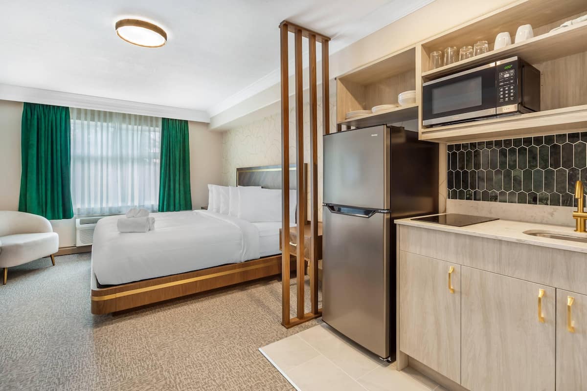 A hotel room with a bed, a kitchenette with a microwave and refrigerator, a wooden partition, and a window with green curtains.
