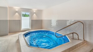 A hot tub with a metal ladder, surrounded by beige and gray tiled walls, with a window and a view of trees. - Velora Hinton, an Ascend Collection Hotel (Hinton)