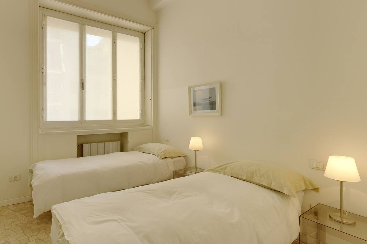 A bedroom with two beds, a window, a radiator, and a wall-mounted picture.