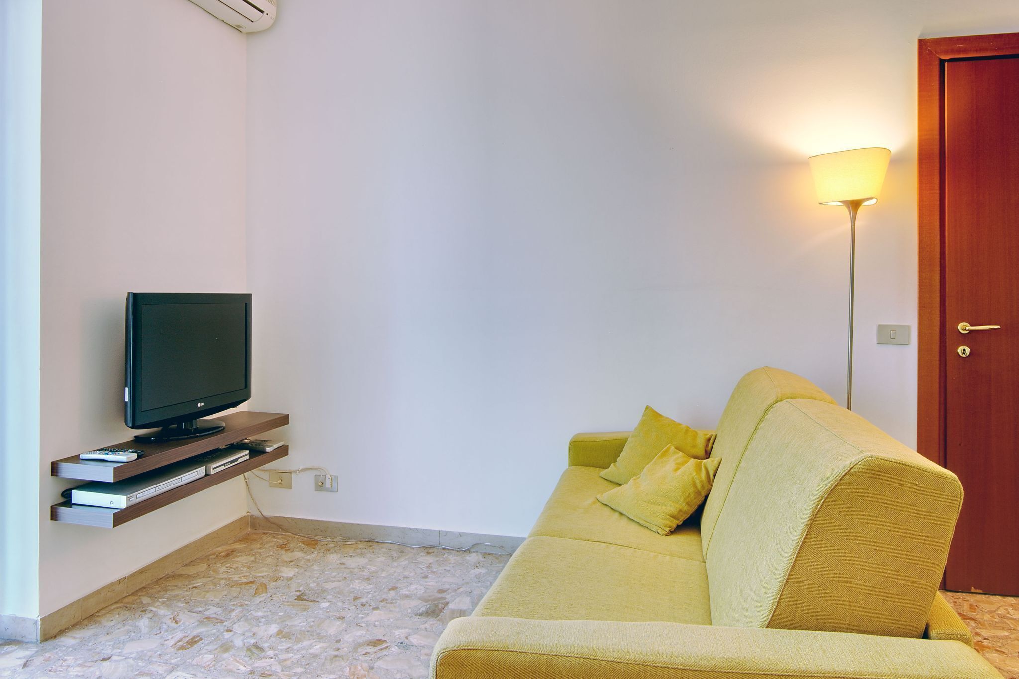 A living room with a flat-screen TV on a stand, a yellow sofa, and a floor lamp.
