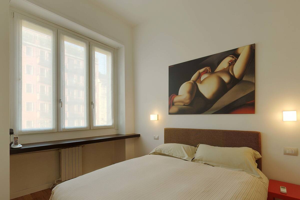 A bedroom with a large bed, a window with blinds, a painting on the wall, and a bedside table with a lamp.