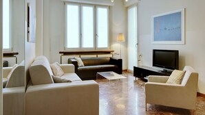 A modern living room with a sofa, armchairs, a coffee table, a TV, and a large window with blinds.