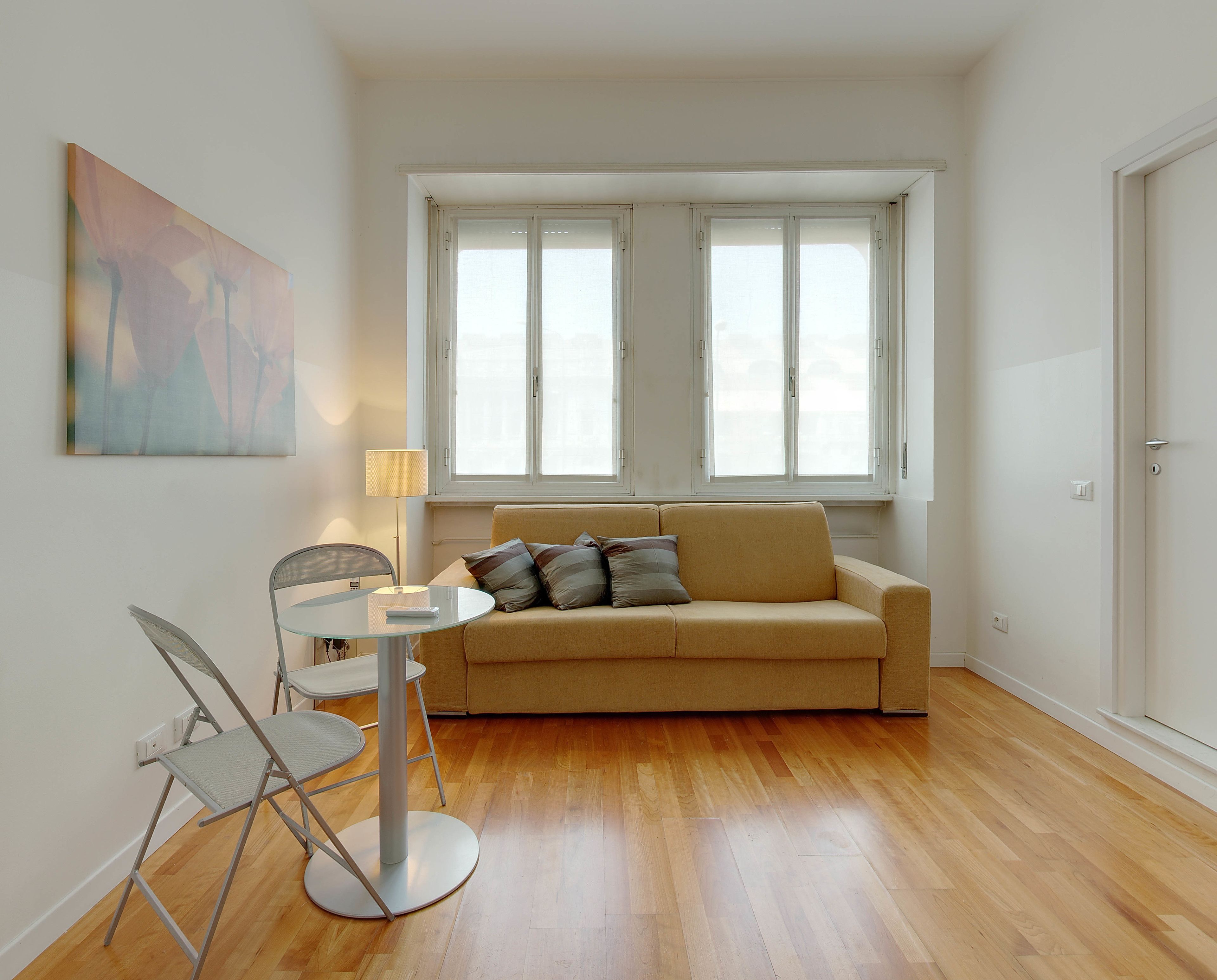 Un salon avec un canapé beige, une table en verre et deux chaises. Il y a un tableau au mur et une fenêtre avec des stores blancs.