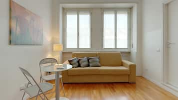 Un salon avec un canapé beige, une table en verre et deux chaises. Il y a un tableau au mur et une fenêtre avec des stores blancs.