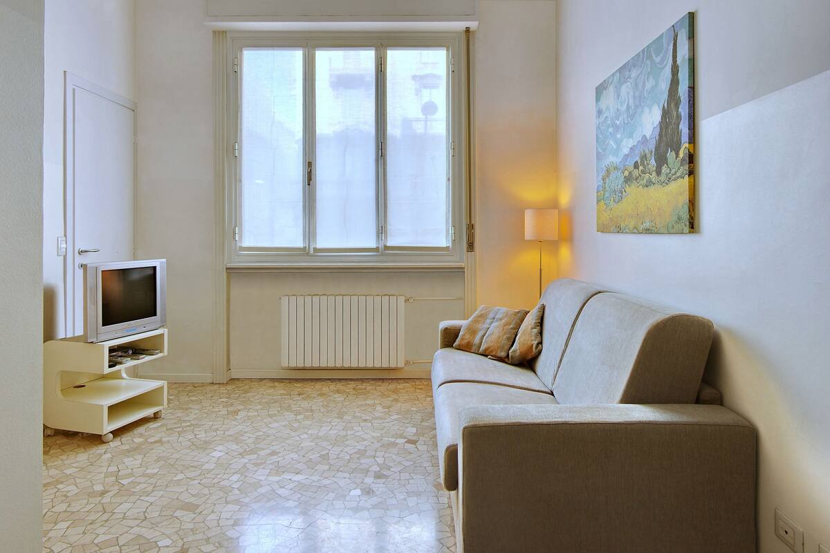 A living room with a sofa, a television on a stand, a window, and a painting on the wall.