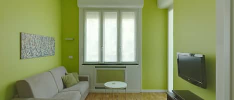 A living room with a sofa, a small round table, a flat-screen TV, and a window with blinds.
