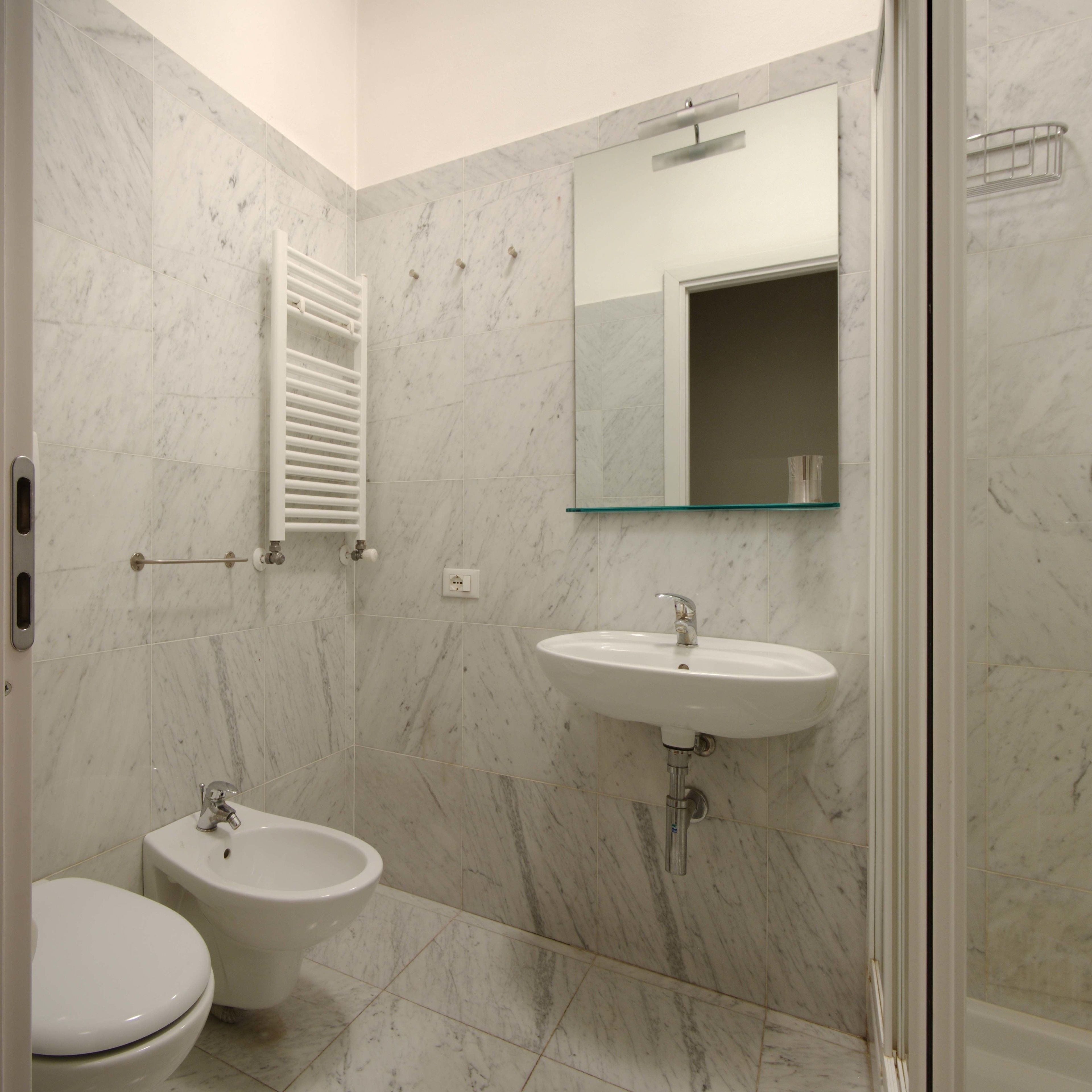 Een badkamer met toilet, bidet en wastafel, alles met marmeren wanden en een glazen plank boven de wastafel.