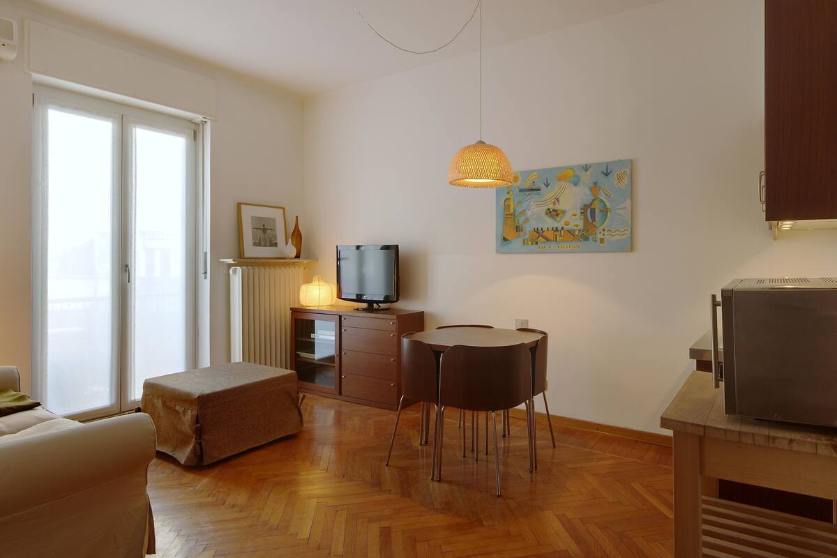 A living room with a sofa, a dining table with chairs, a television, a radiator, and a painting on the wall.
