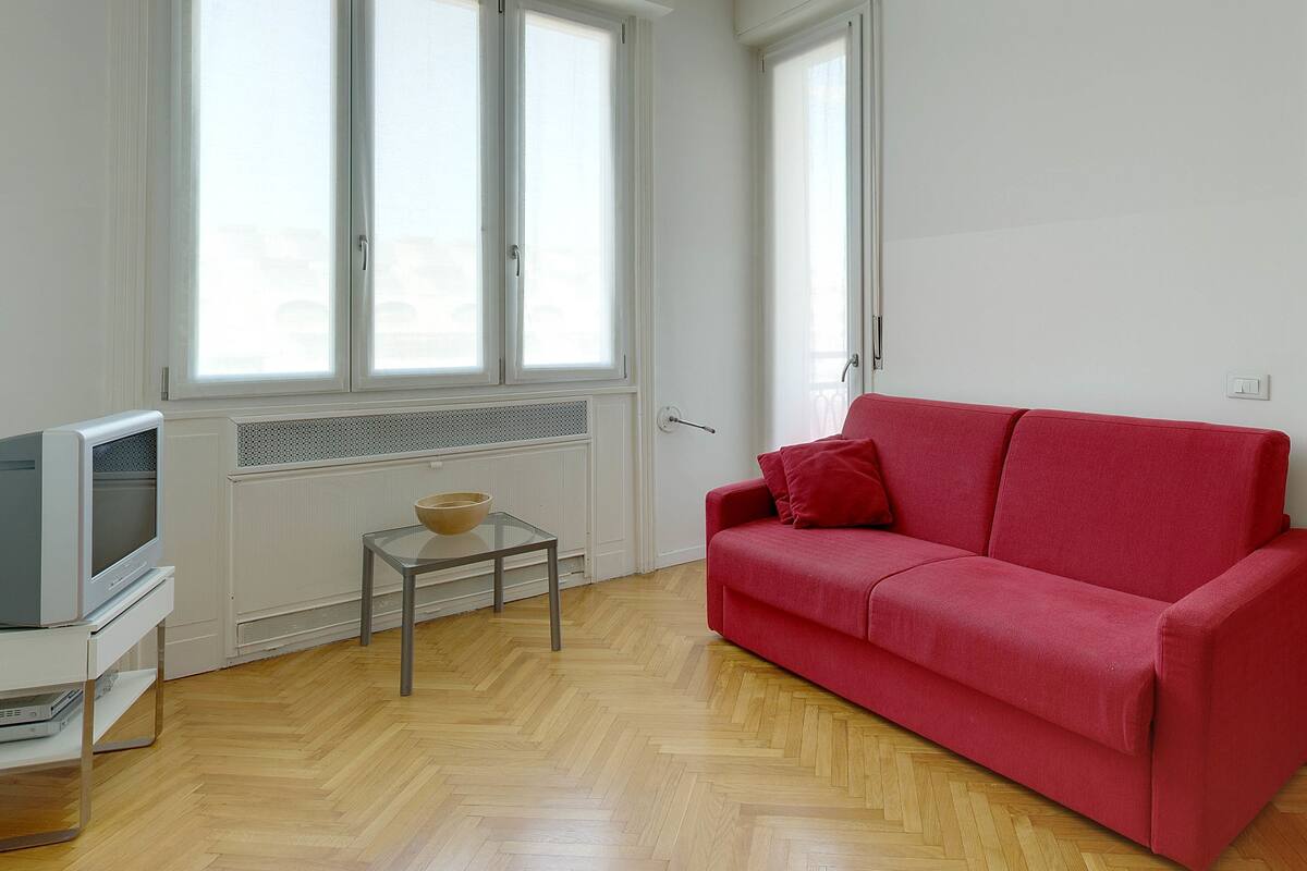 A room with a red sofa, a small glass table with a bowl, a television, and large windows with white frames.