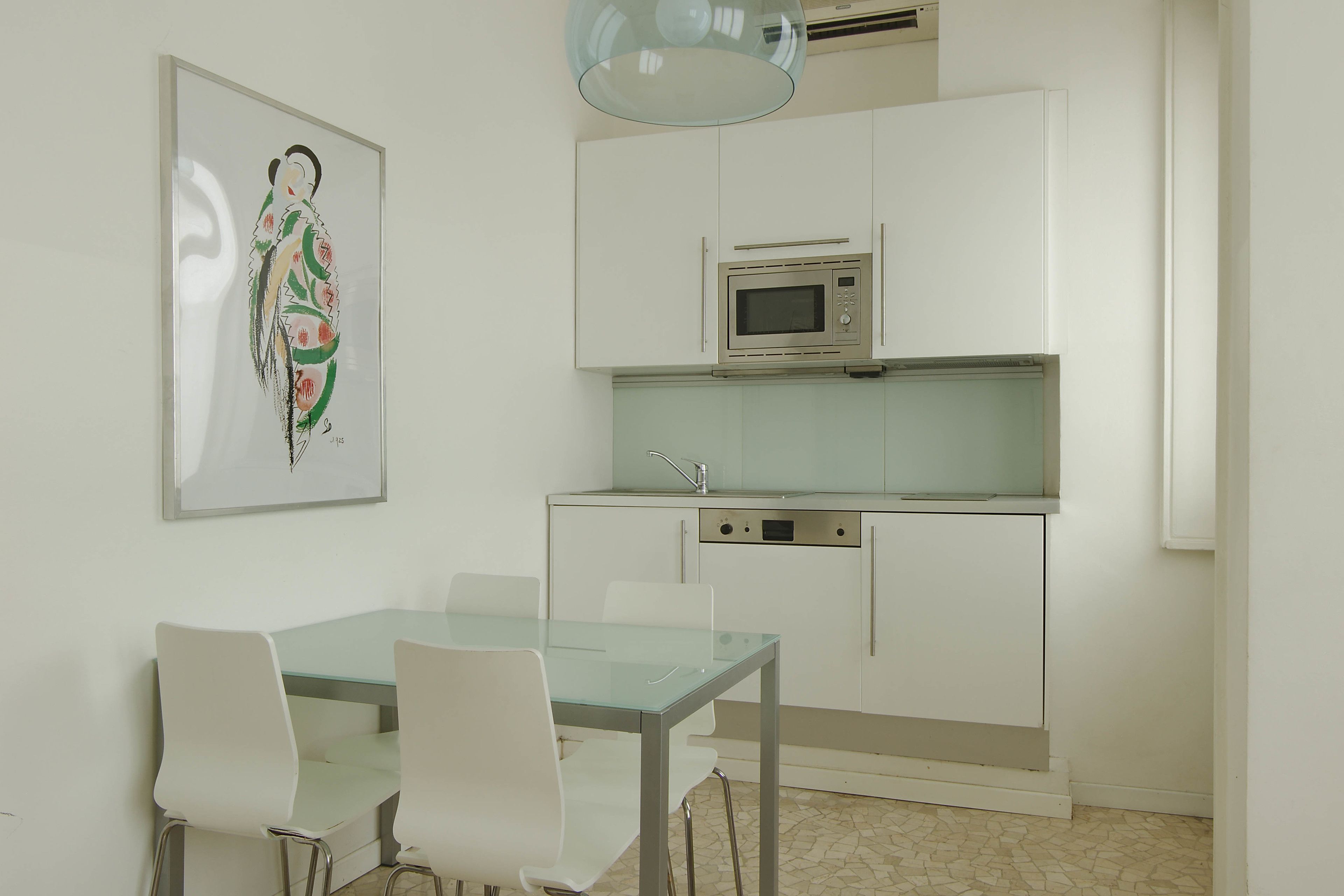 A modern kitchen with white cabinets, a stainless steel microwave, and a dishwasher. There is a glass table with four white chairs. A framed artwork is visible on the wall.