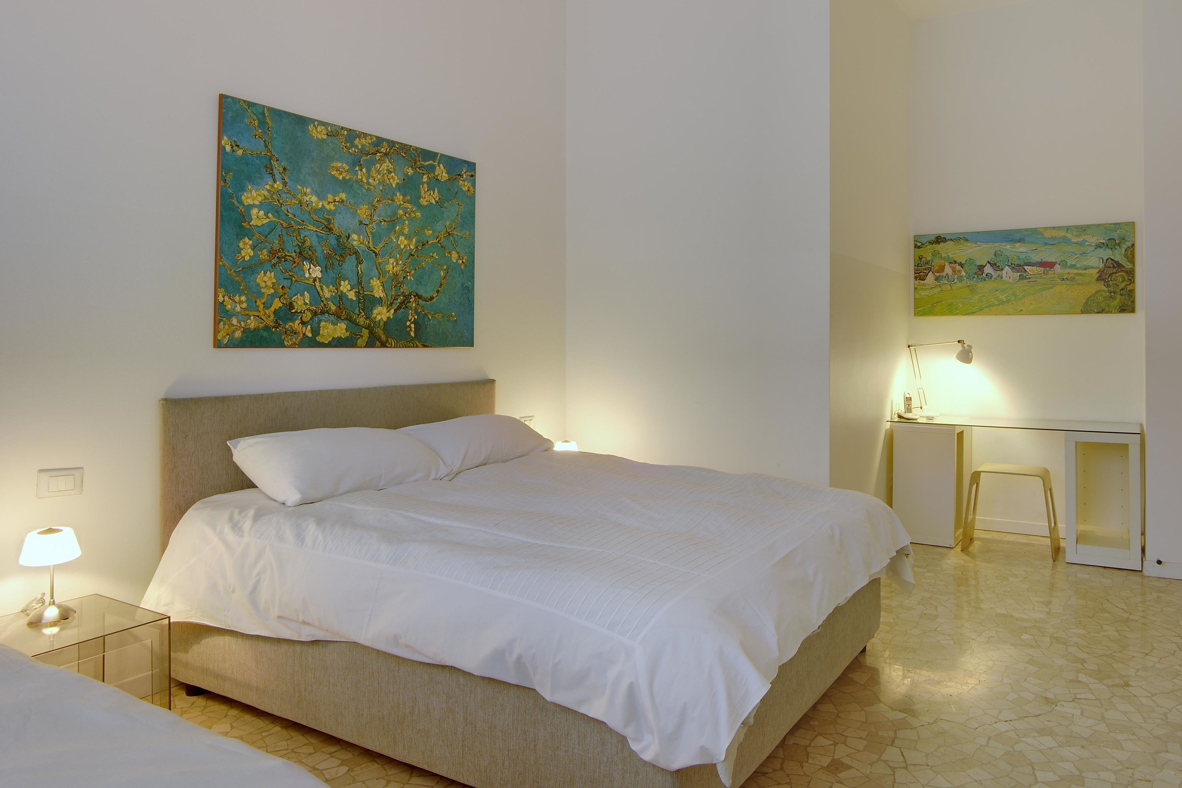 A bedroom with a bed, a nightstand, a desk, and two paintings on the walls.