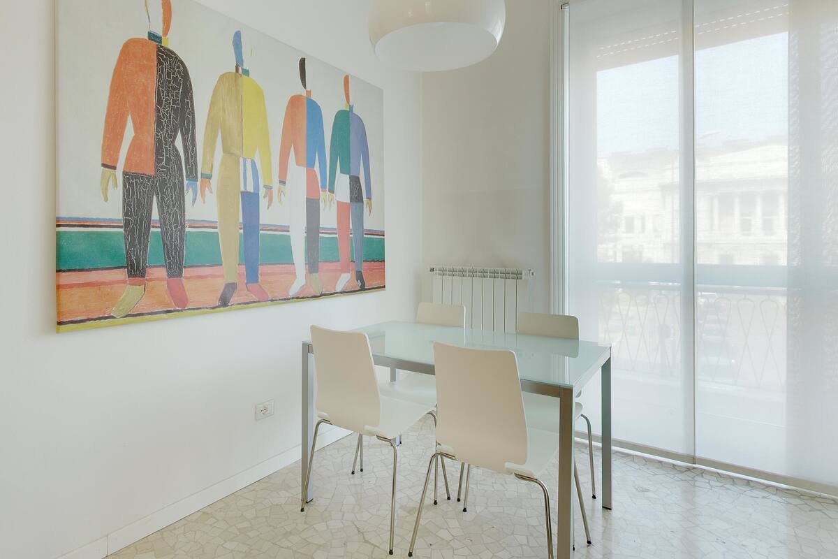 A modern dining area with a glass table and chairs, a large abstract painting on the wall, and a large window with a view of buildings.