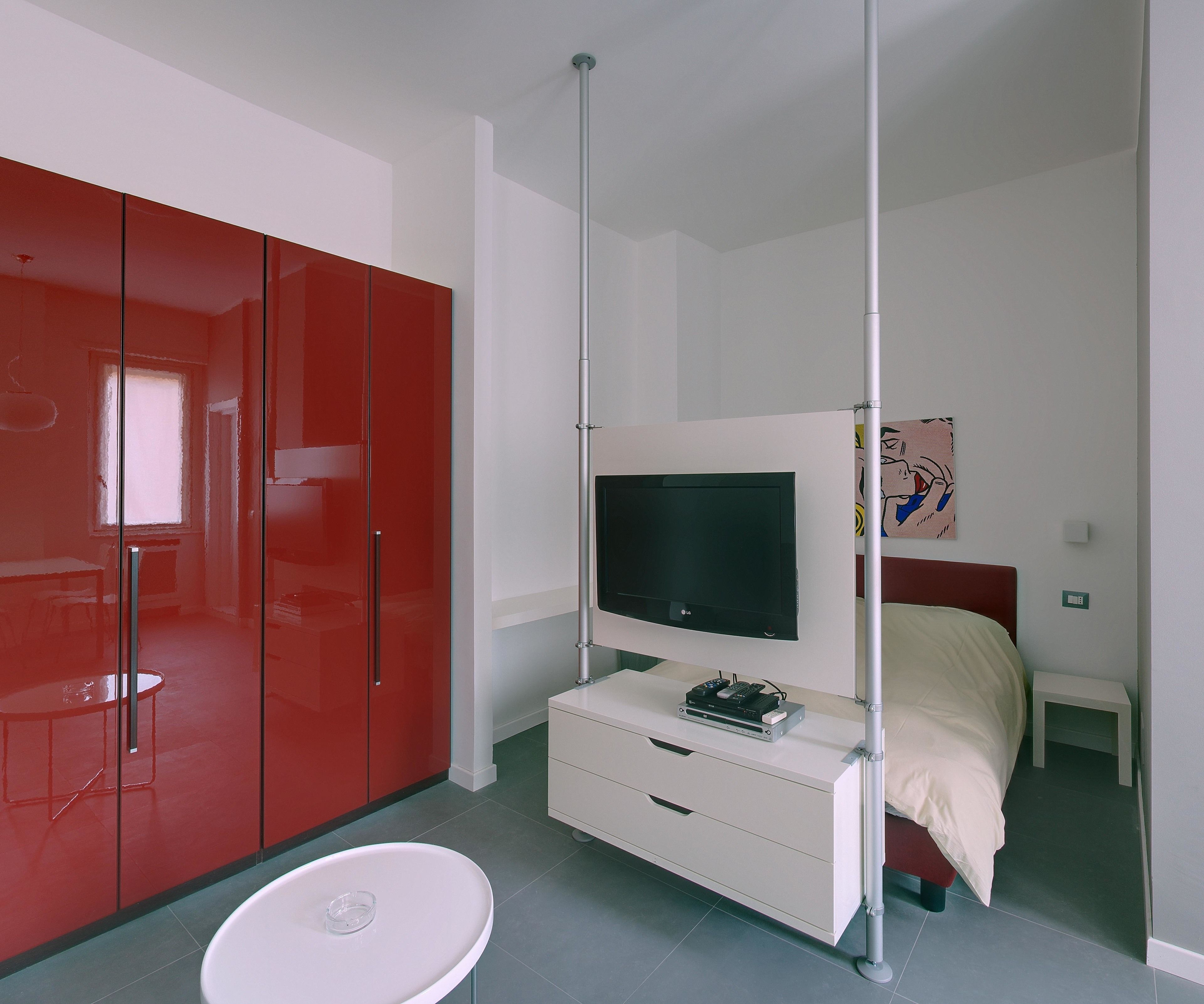 A modern bedroom with a bed, a TV mounted on a white dresser, a red wardrobe, and a small white table.