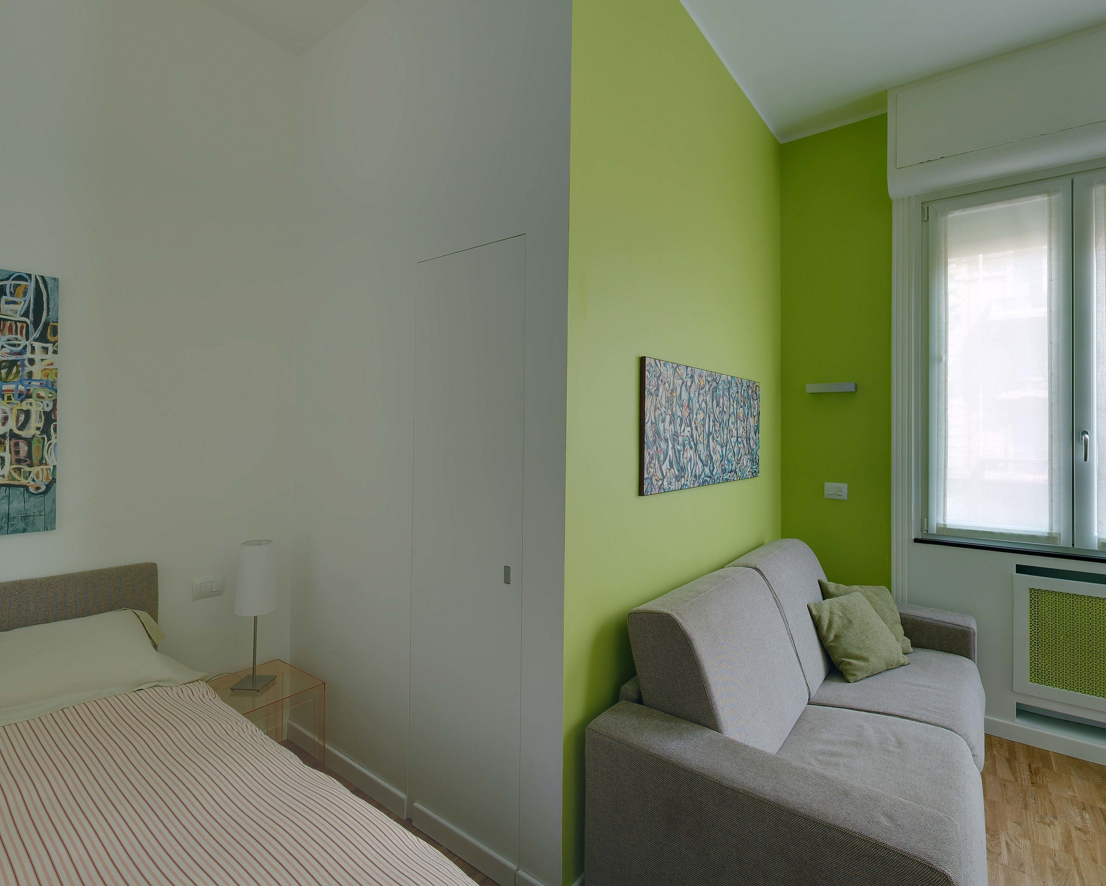 A room with a bed, a nightstand, a lamp, a sofa, a green accent wall, and a window with blinds.