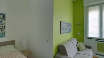 A room with a bed, a nightstand, a lamp, a sofa, a green accent wall, and a window with blinds.