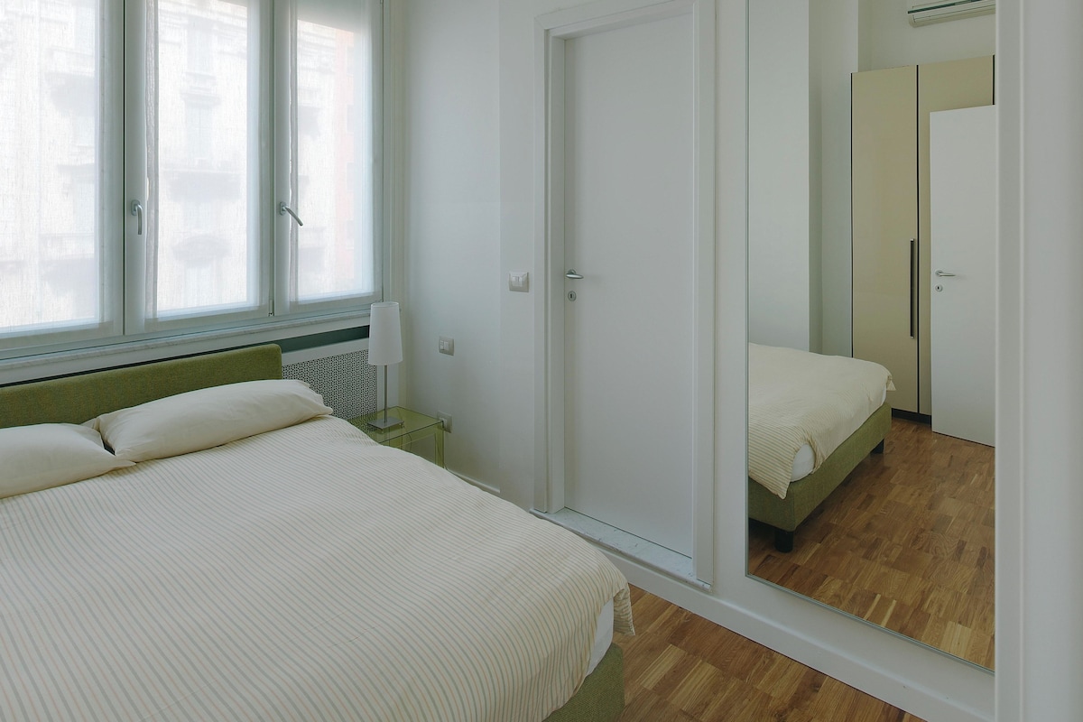 A bedroom with a bed, a nightstand, a lamp, a wardrobe, and a mirror.