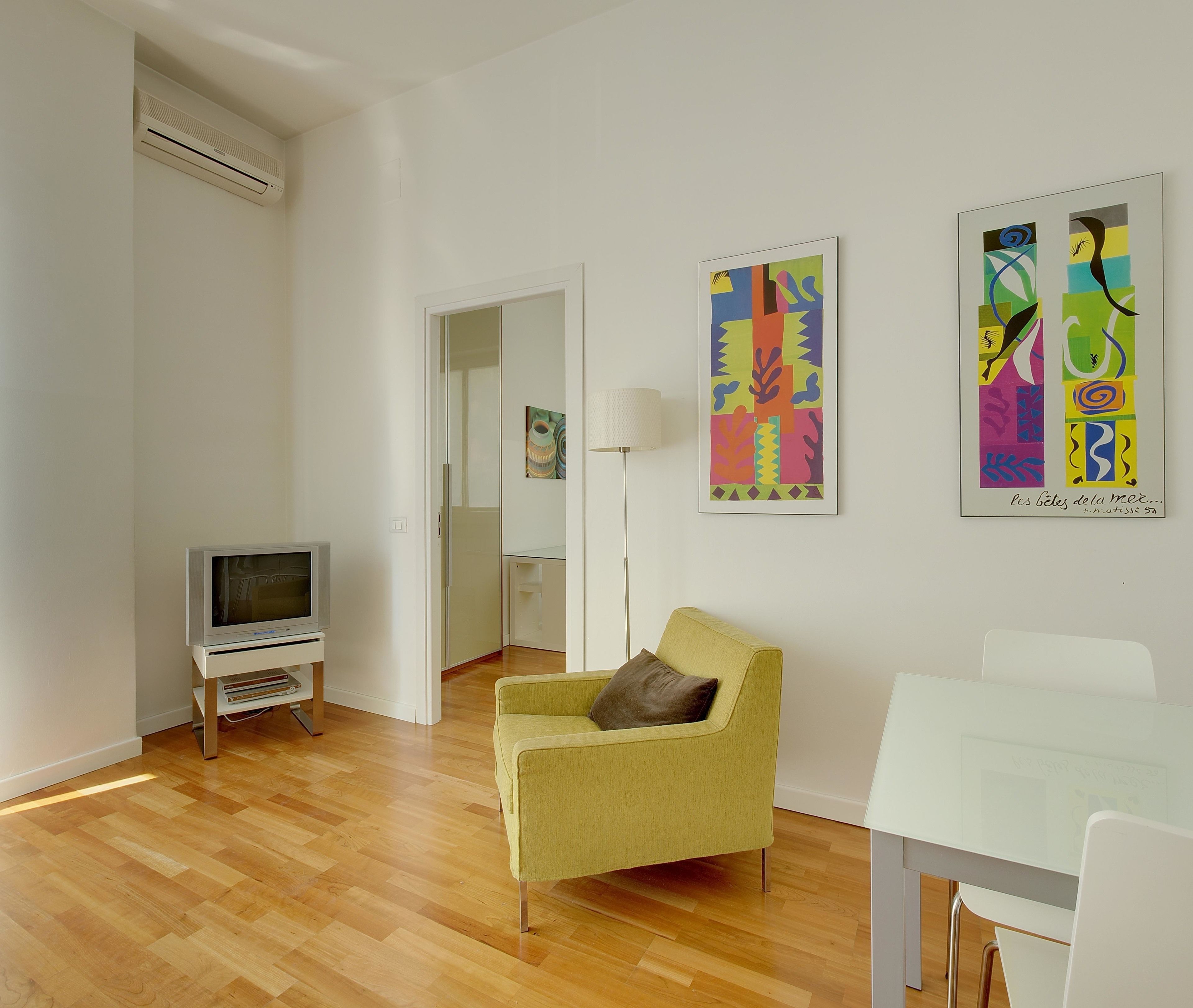 A modern living room with a flat-screen TV, a yellow armchair, a white dining table, and abstract art on the walls.