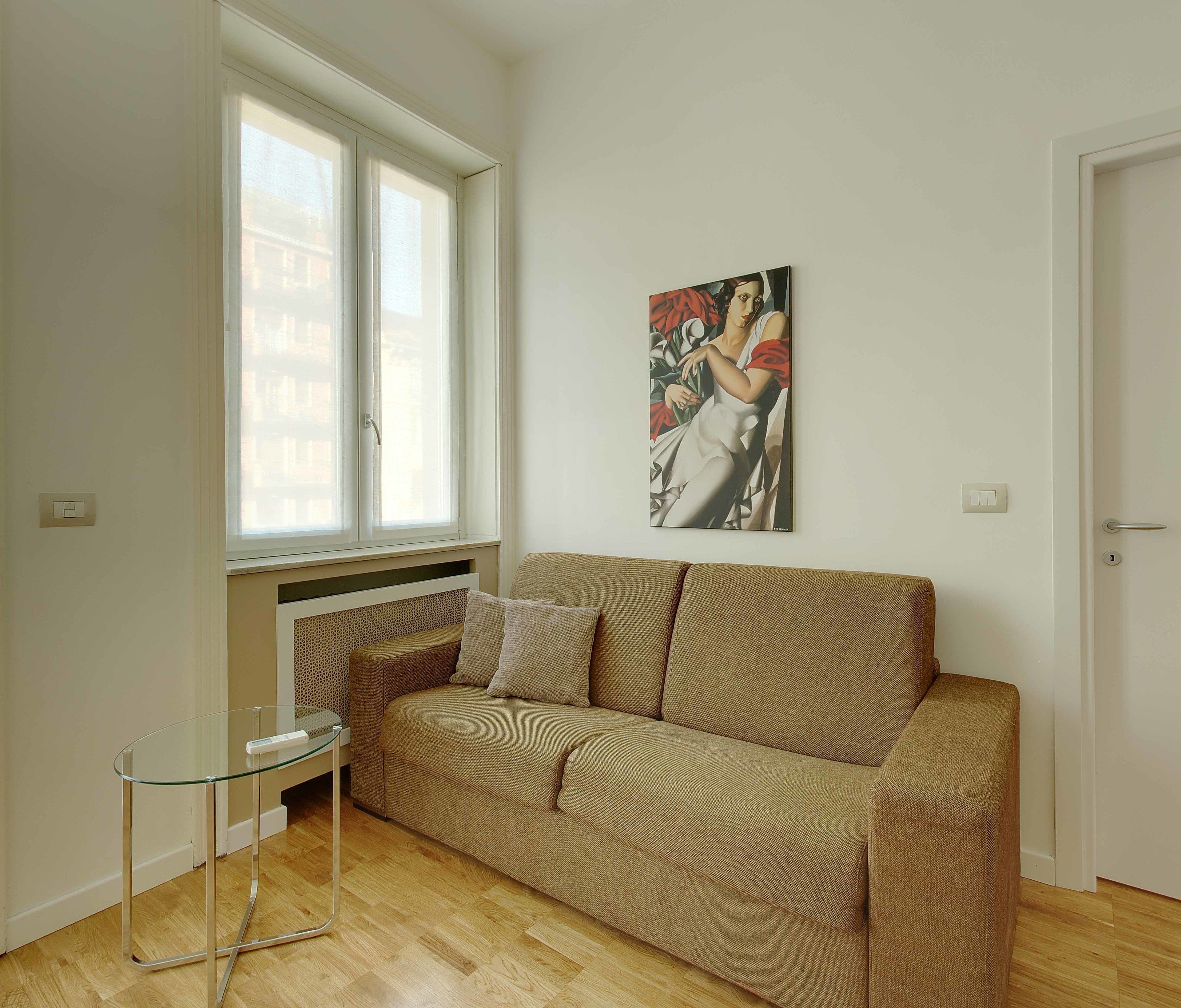 Una habitación con un sofá color crema, una mesa de centro de vidrio, una ventana con persianas y un cuadro en la pared.