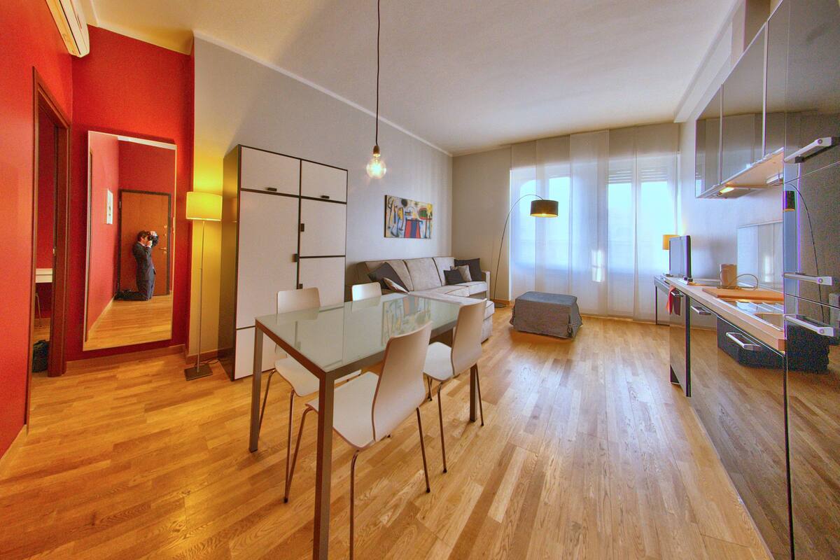 A modern living room with a glass dining table, a sofa, and a large window.
