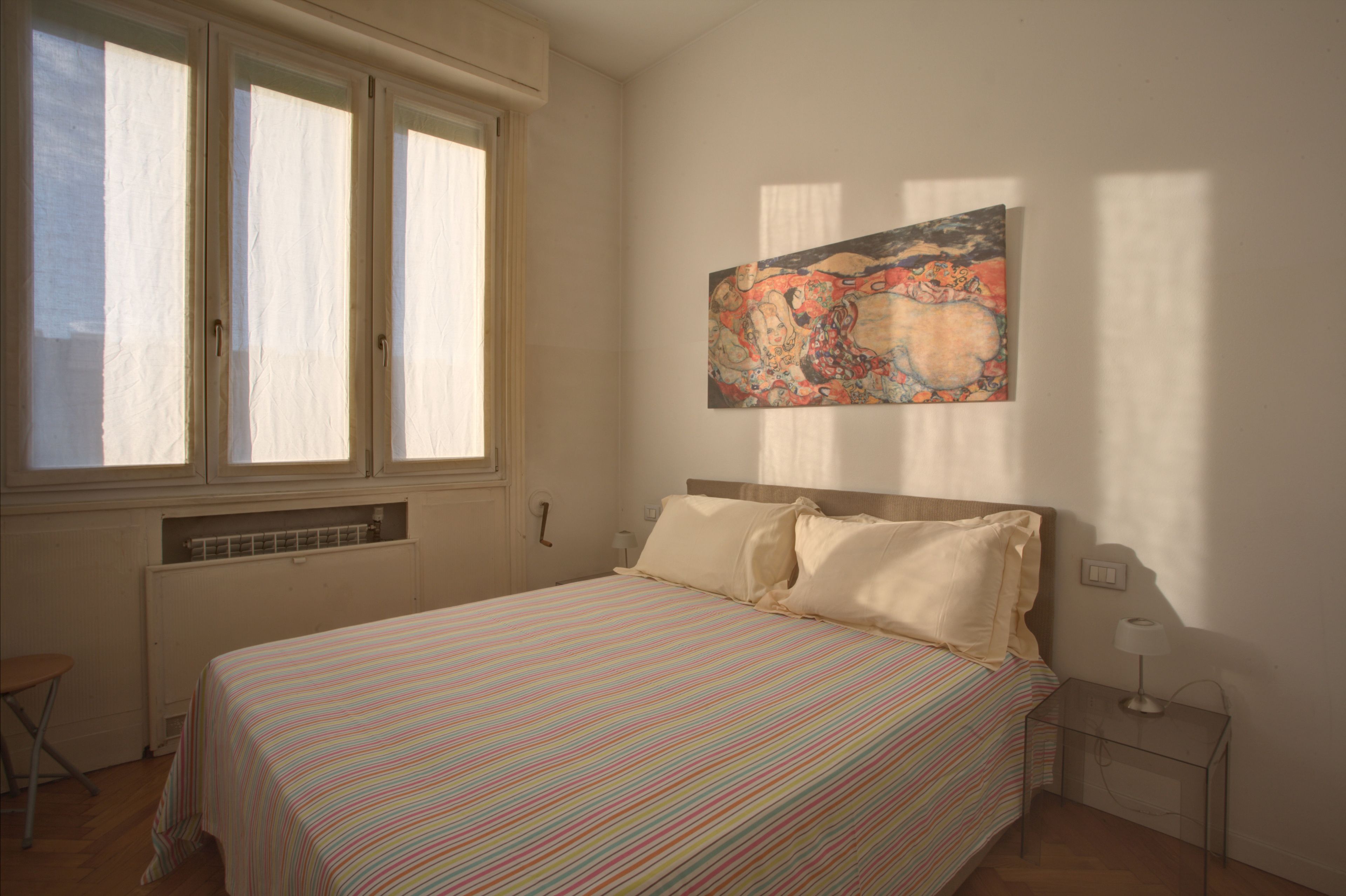 A bedroom with a bed, a window, a radiator, a chair, and a painting on the wall.