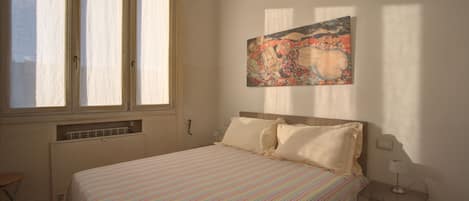 A bedroom with a bed, a window, a radiator, a chair, and a painting on the wall.