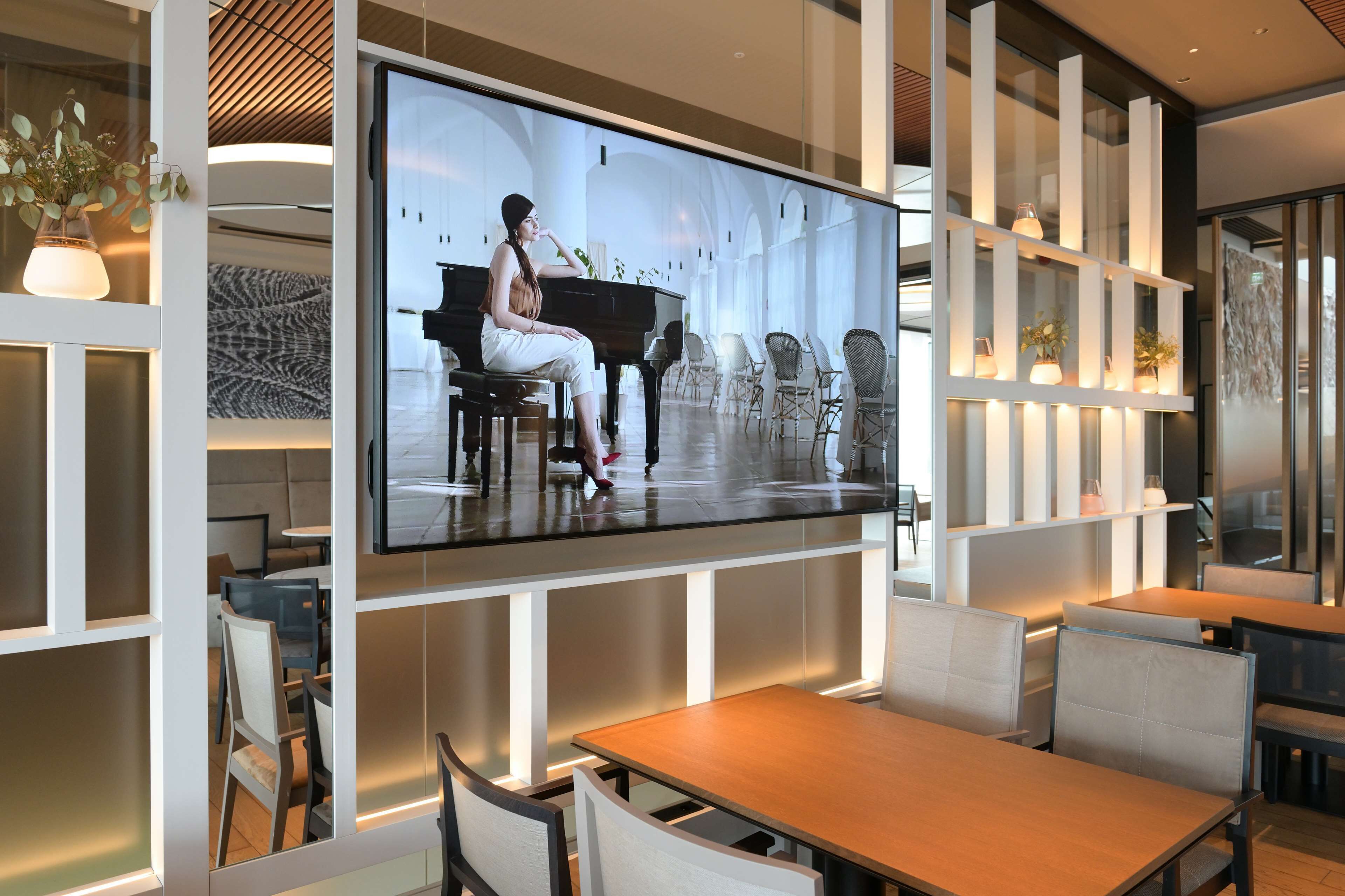 En modern restaurang med en stor TV-skärm som visar en pianist, träbord och stolar.