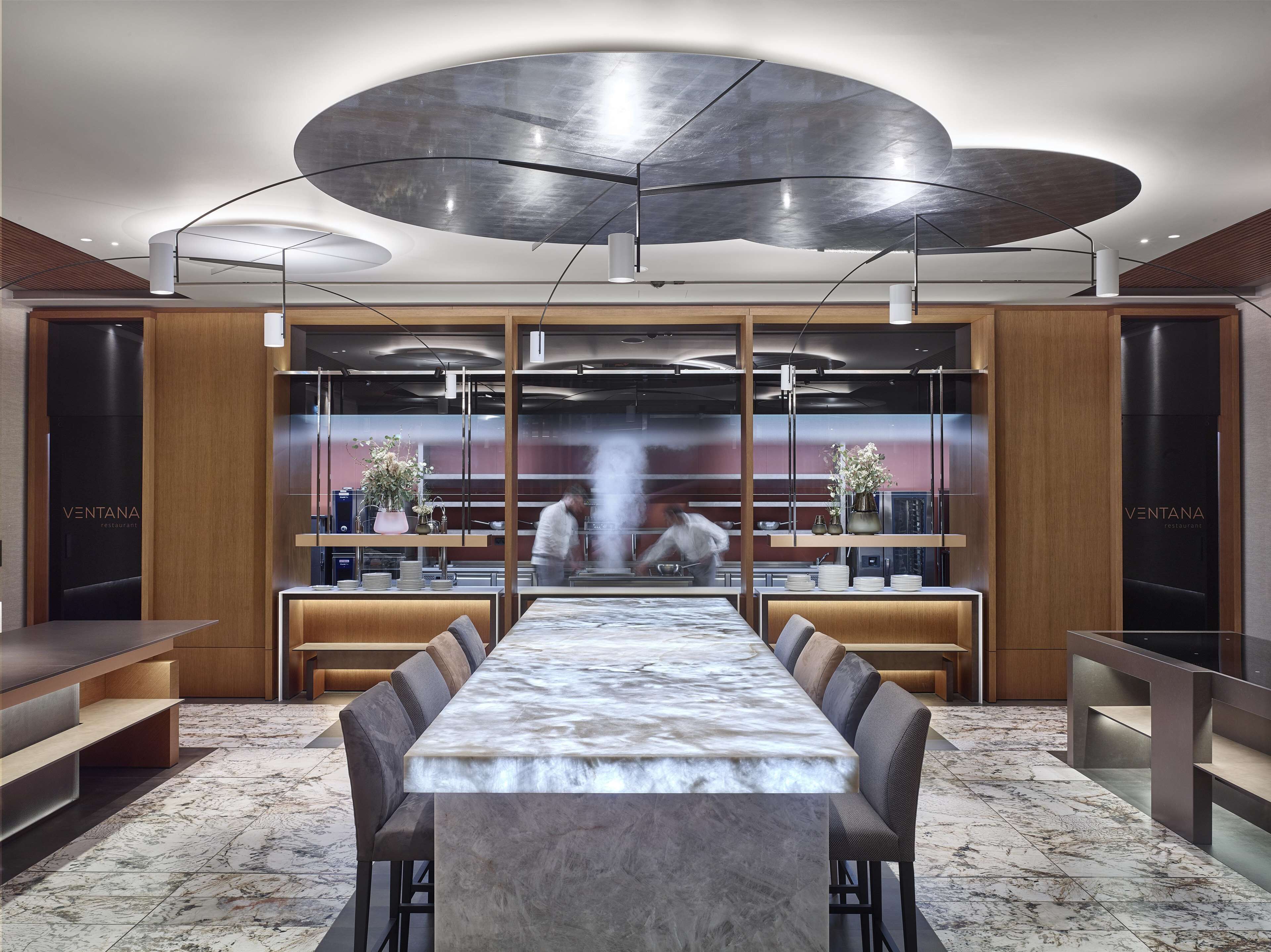 A modern restaurant interior with a long marble table, wooden accents, and a contemporary ceiling design.