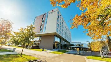 Un hotel moderno con una entrada destacada, un área de estacionamiento y follaje circundante.