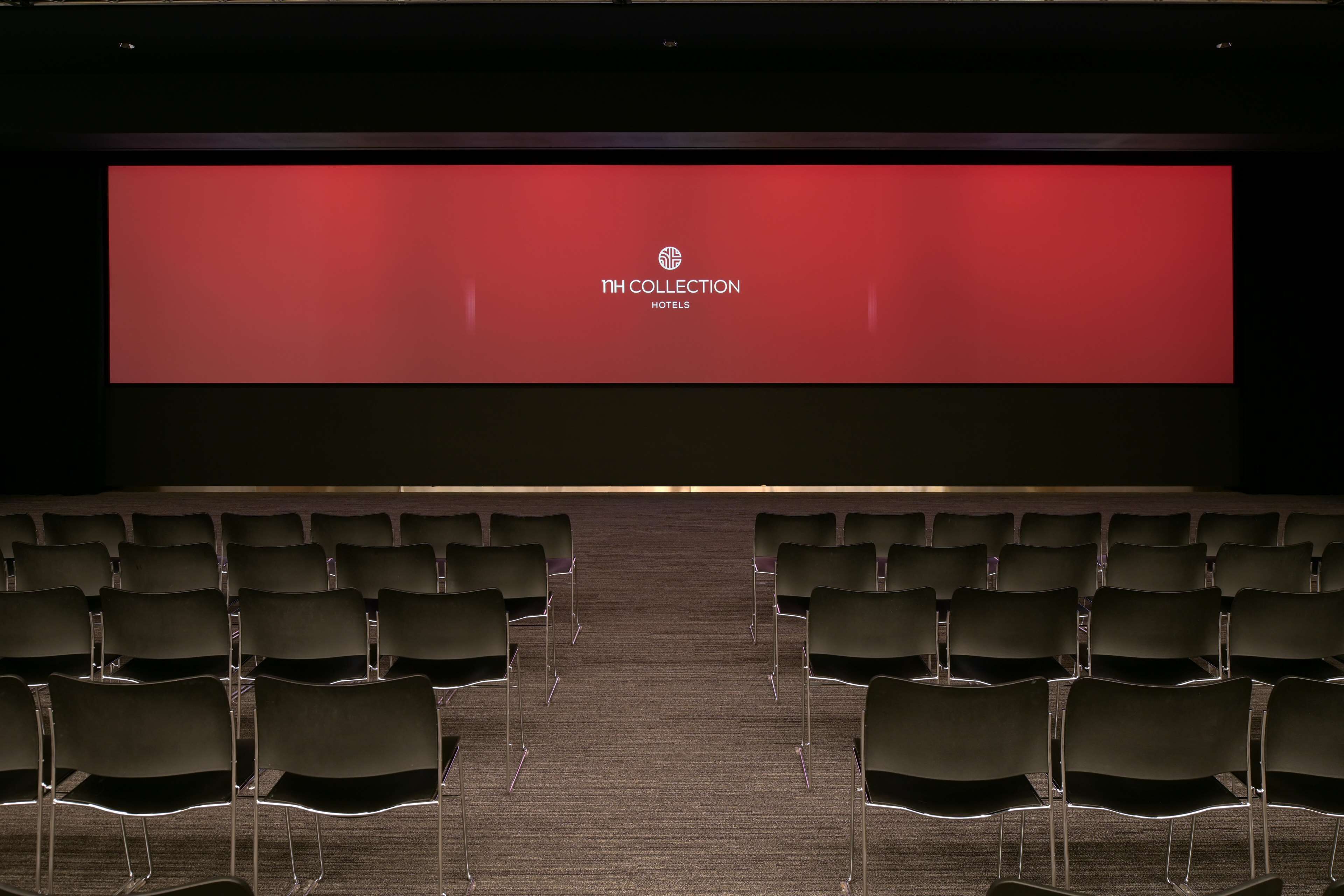 An empty auditorium with rows of chairs facing a large screen displaying "NH COLLECTION HOTELS".