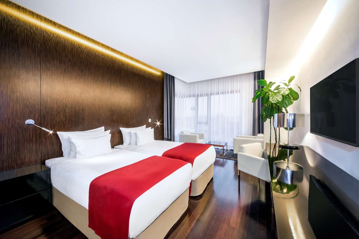 A modern hotel room with a large bed, a TV, a plant, and a desk with a chair.