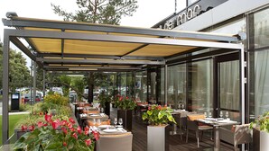 Un espace extérieur aménagé pour les repas, avec des tables et des chaises, protégé par un auvent et entouré de plantes en pot.