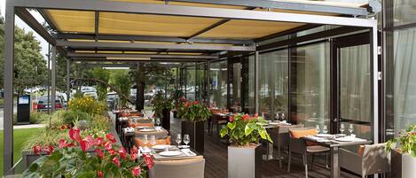 An outdoor dining area with tables and chairs, covered by a canopy, and surrounded by potted plants.