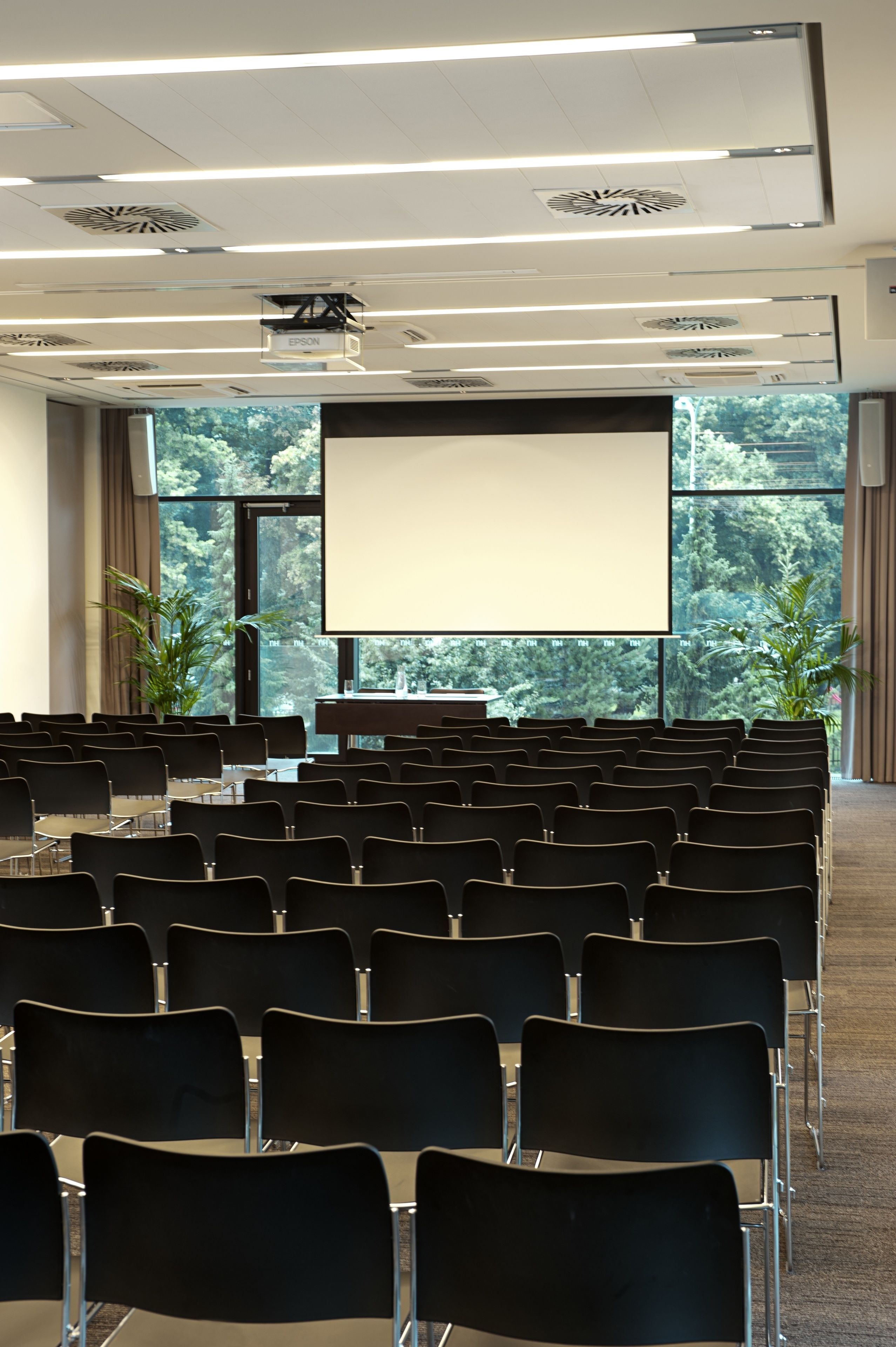 Ein Konferenzraum mit Reihen von Stühlen, einer Projektionsleinwand und Blick auf Bäume durch große Fenster.