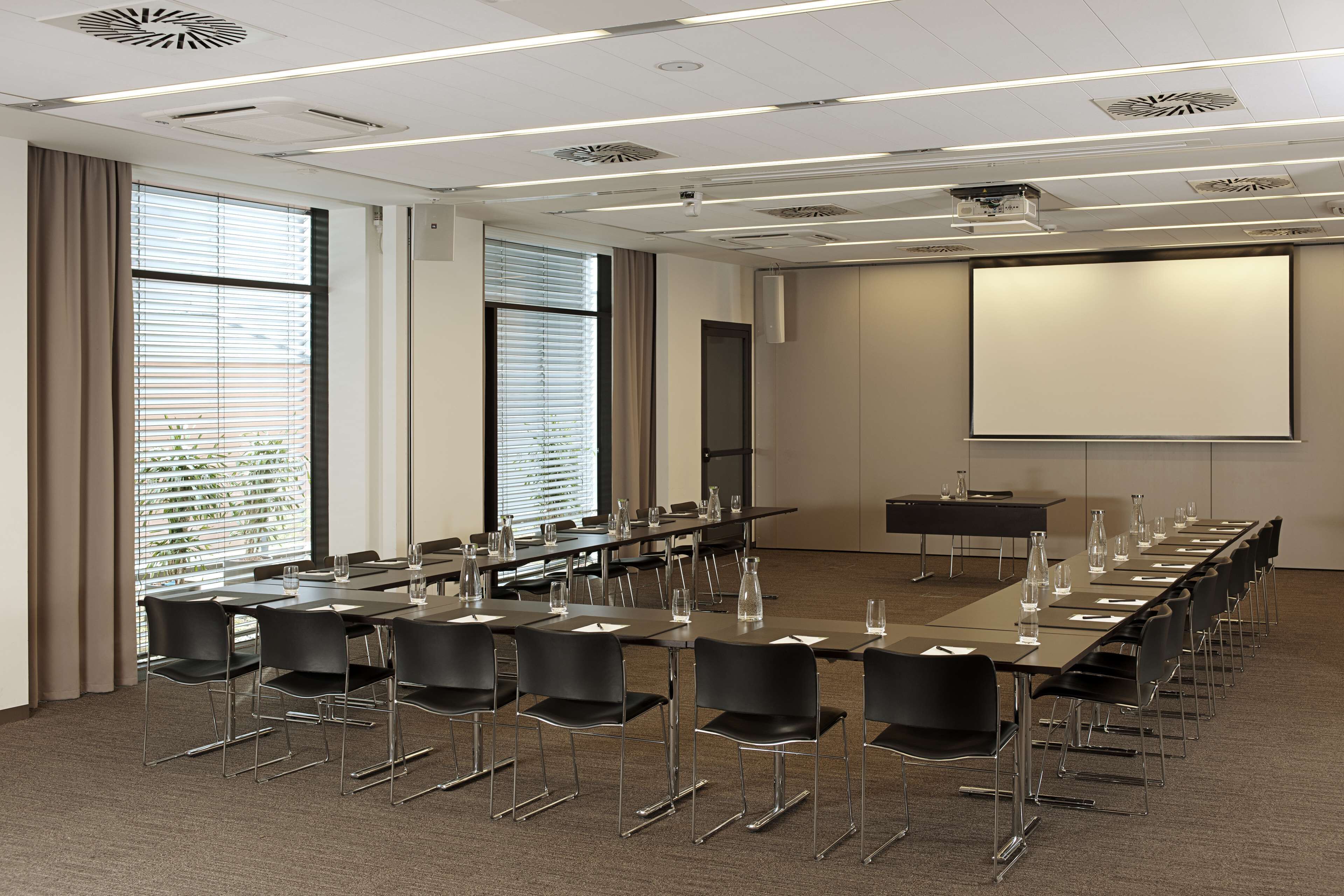 A conference room with a long table, chairs, a projector screen, and large windows with blinds.