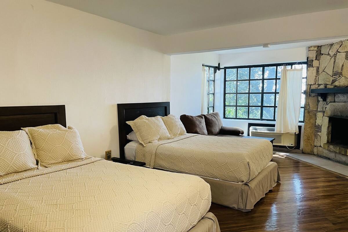 A hotel room with two beds, a stone fireplace, a large window with curtains, and a wooden floor.