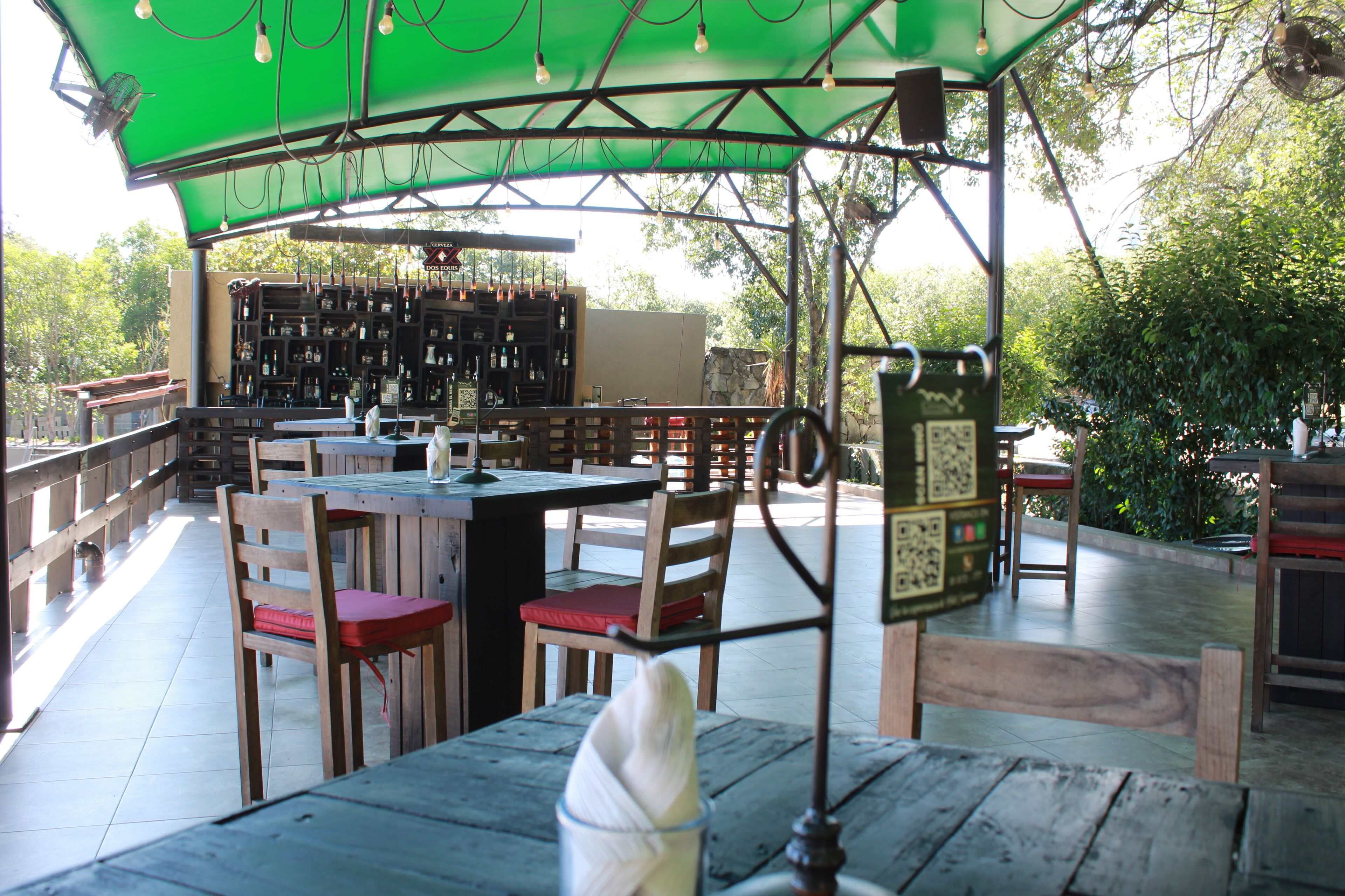 An outdoor dining area with wooden tables and chairs, a bar area with shelves of bottles, and a green canopy overhead.