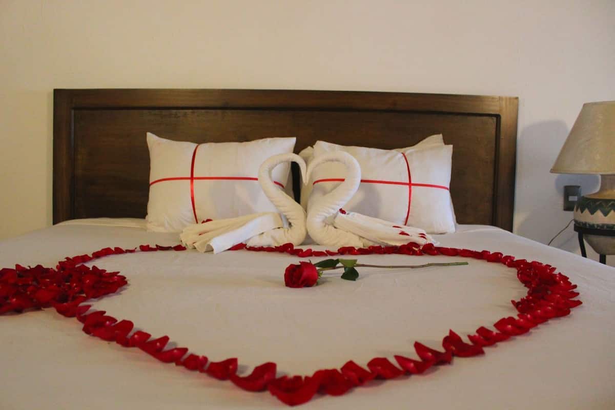 A bed with a heart-shaped arrangement of rose petals and two towels shaped like swans tied together.