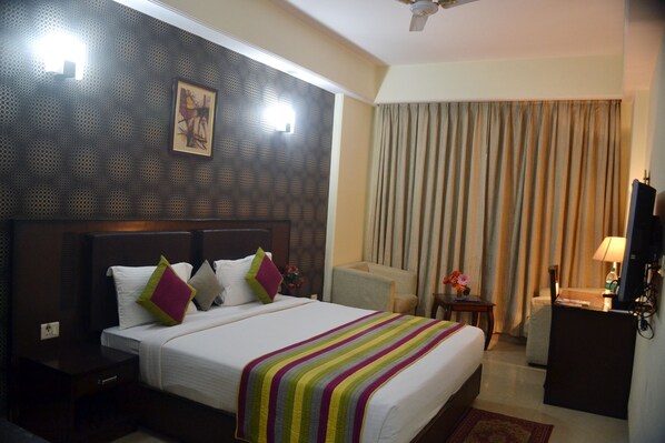 A hotel room with a large bed, a headboard with a patterned design, a flat-screen TV, a desk with a lamp, and a window with curtains. - Hotel Venus (New Delhi)